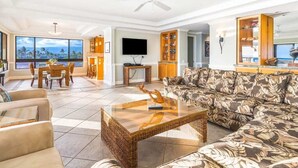 Un vaste salon comprenant un grand canapé, une table basse et un coin repas avec une table et des chaises. Un téléviseur est fixé au mur et un ventilateur de plafond est installé.