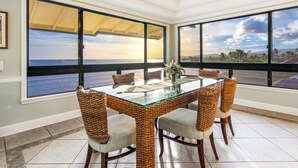 Un coin repas avec une table en verre entourée de chaises en osier, de grandes fenêtres offrant une vue sur l’océan et un luminaire suspendu au plafond.