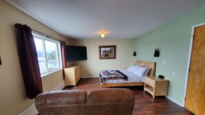 A bedroom with a bed, a TV, a dresser, and a window with curtains. - {Forget-Me-Not Inn} Alaskan Waterfront Vacation Rental (Deer Suite) (Sitka)