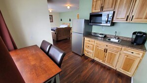 A compact kitchen with wooden cabinets, a microwave, a refrigerator, and a dining area with a table and chairs. - {Forget-Me-Not Inn} Alaskan Waterfront Vacation Rental (Deer Suite) (Sitka)