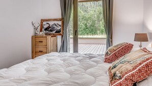 A bedroom with a bed, a dresser, a framed picture, a lamp, and a view of a forest through a window.