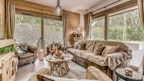 A cozy cabin interior with a wooden ceiling, large windows, a sofa, a coffee table, and a fireplace.