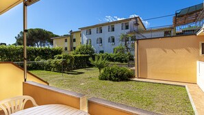En balkon med et bord og stole, en lille have med buske og græsplæne, samt en bygning med flere vinduer og balkoner i baggrunden.