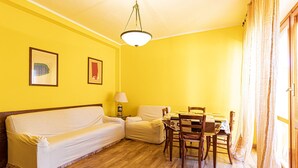 Et værelse med gul væg, trægulv, en dækket sofa, et bord med stole og en hængende lampe.