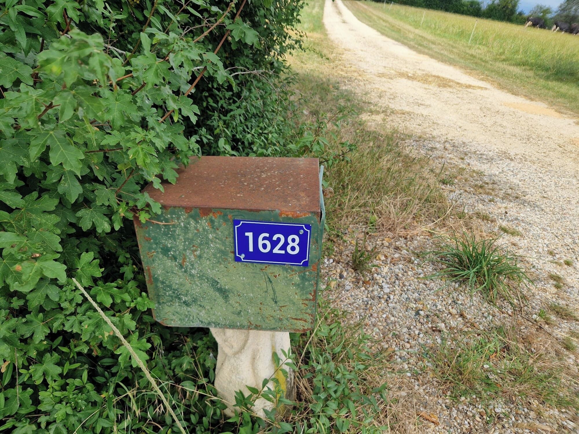 A rustic mailbox with a blue sign displaying the number 1628, situated beside a gravel path and surrounded by greenery.