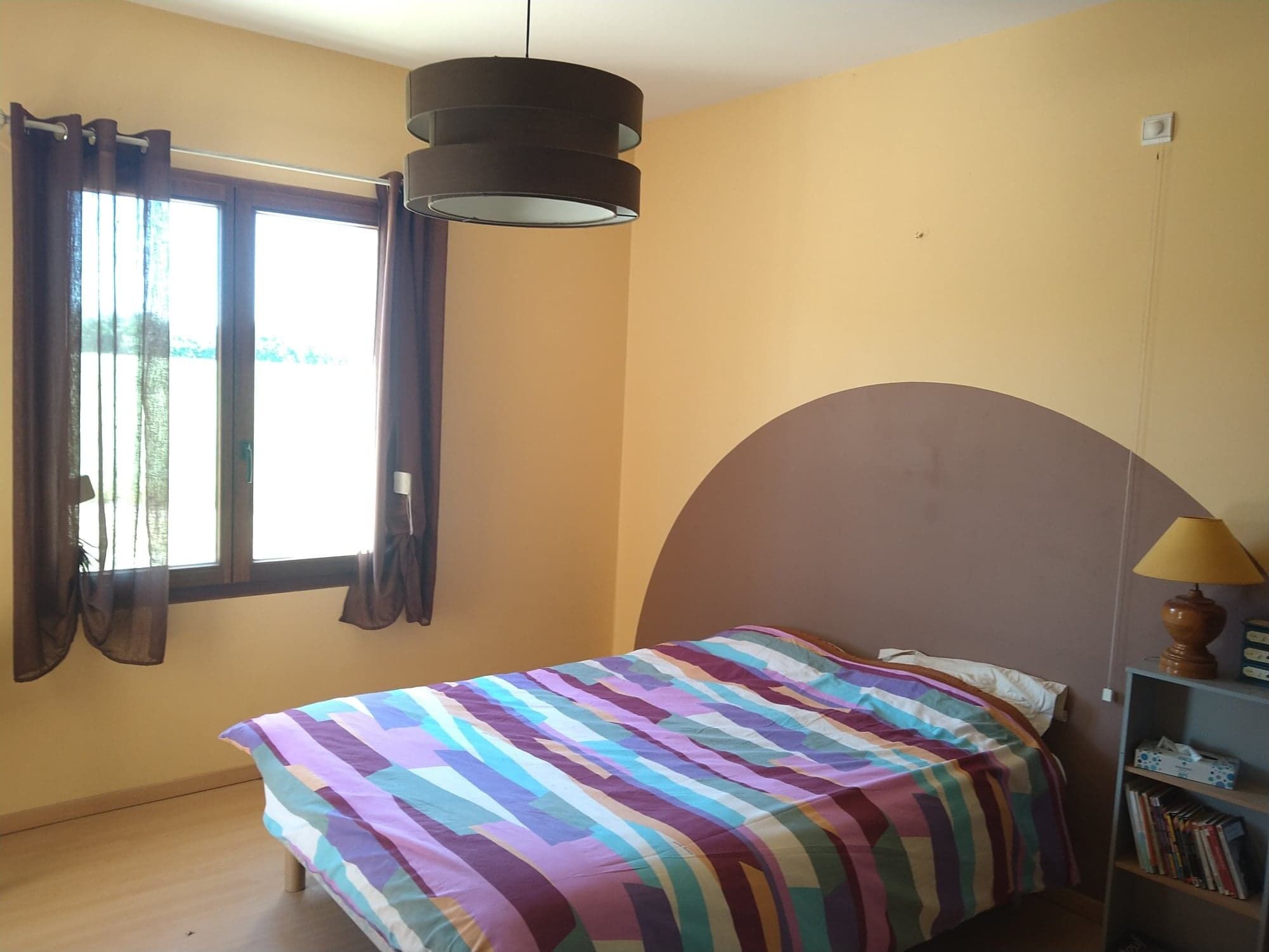 A bedroom with a bed, a window with curtains, a bookshelf, and a hanging lamp.