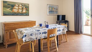 A dining area with a table covered in a fish-patterned tablecloth, wooden chairs, a wooden cabinet, a television, and a painting of a sailing ship. - APPARTAMENTI SECCHETO - BELLAVISTA (Campo nell'Elba)