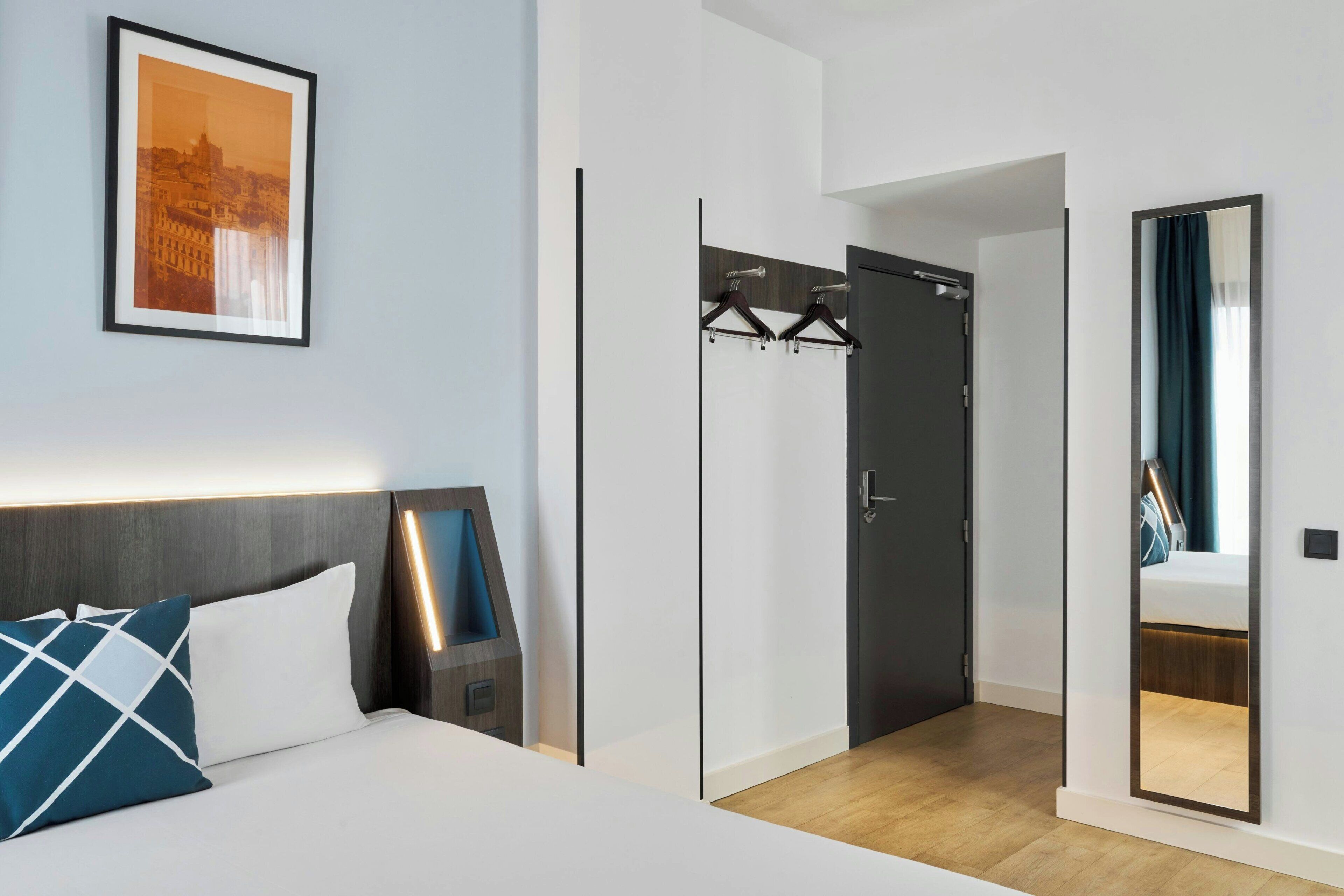 A modern hotel room with a bed, a nightstand, a framed picture, a coat rack, and a mirror.