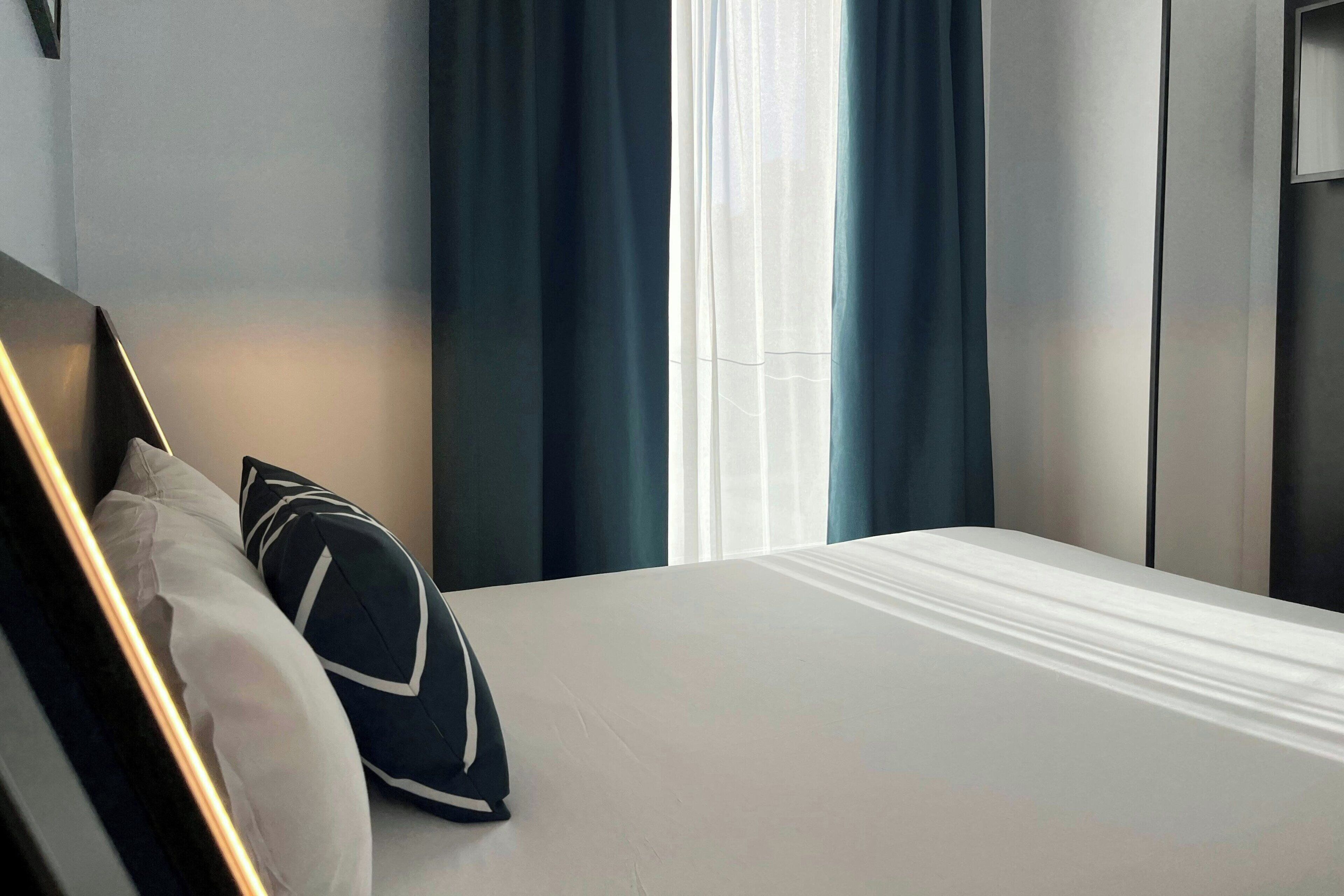 Ein ordentlich bezogenes Bett mit weißen Leinen und zwei Kissen, ein Fenster mit blauen Vorhängen und ein modernes Wanddesign.