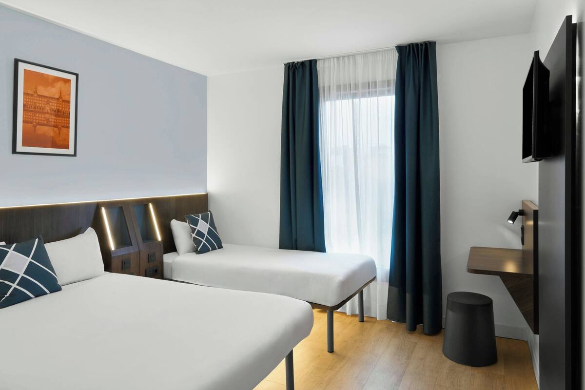 A hotel room with two beds, a TV, a small table, a trash bin, and a window with curtains.