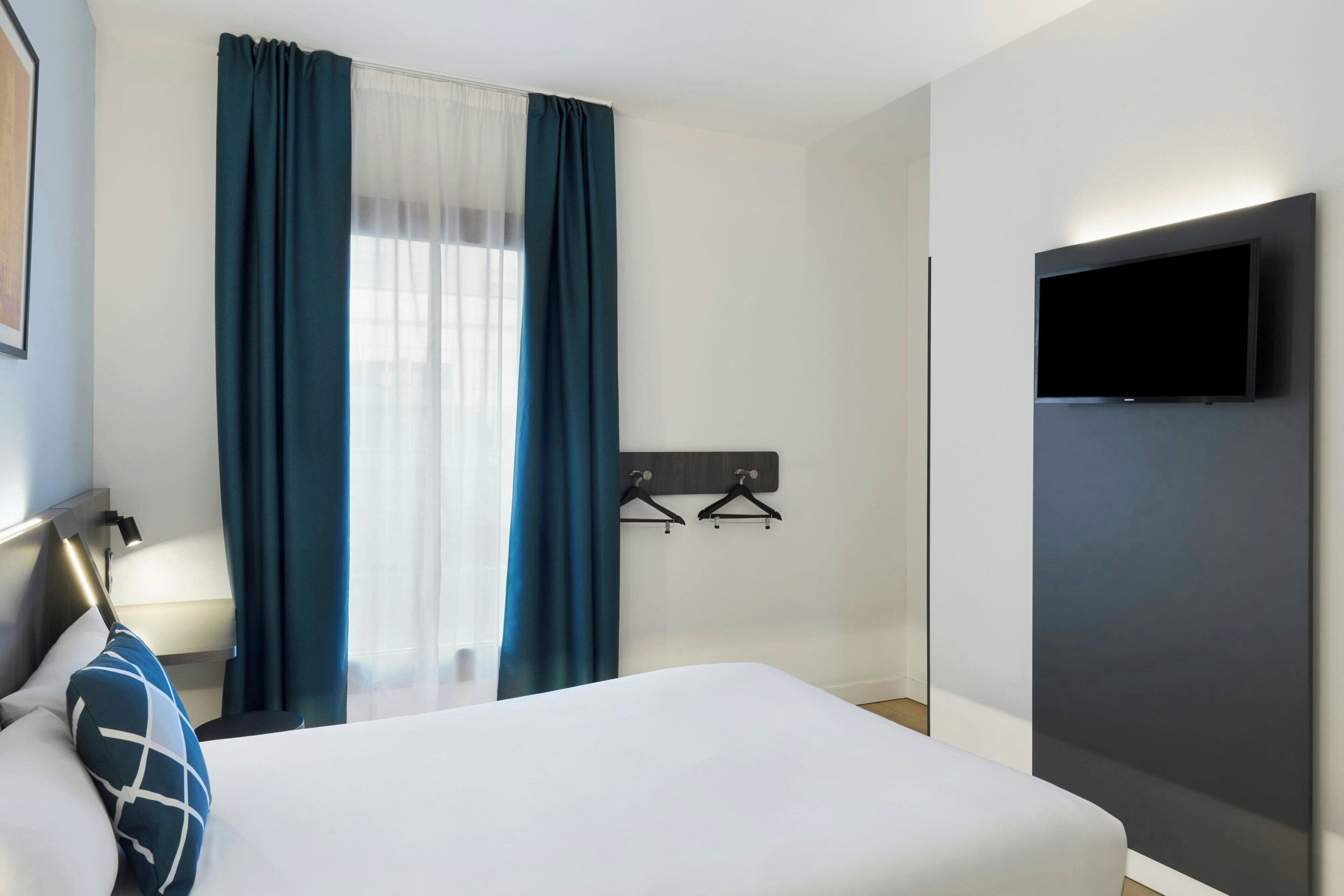 Een hotelkamer met een bed, een aan de muur bevestigde televisie en een raam met gordijnen.