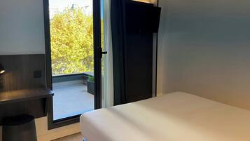 A modern hotel room with a bed, a desk, and a large window offering a view of trees and a balcony.