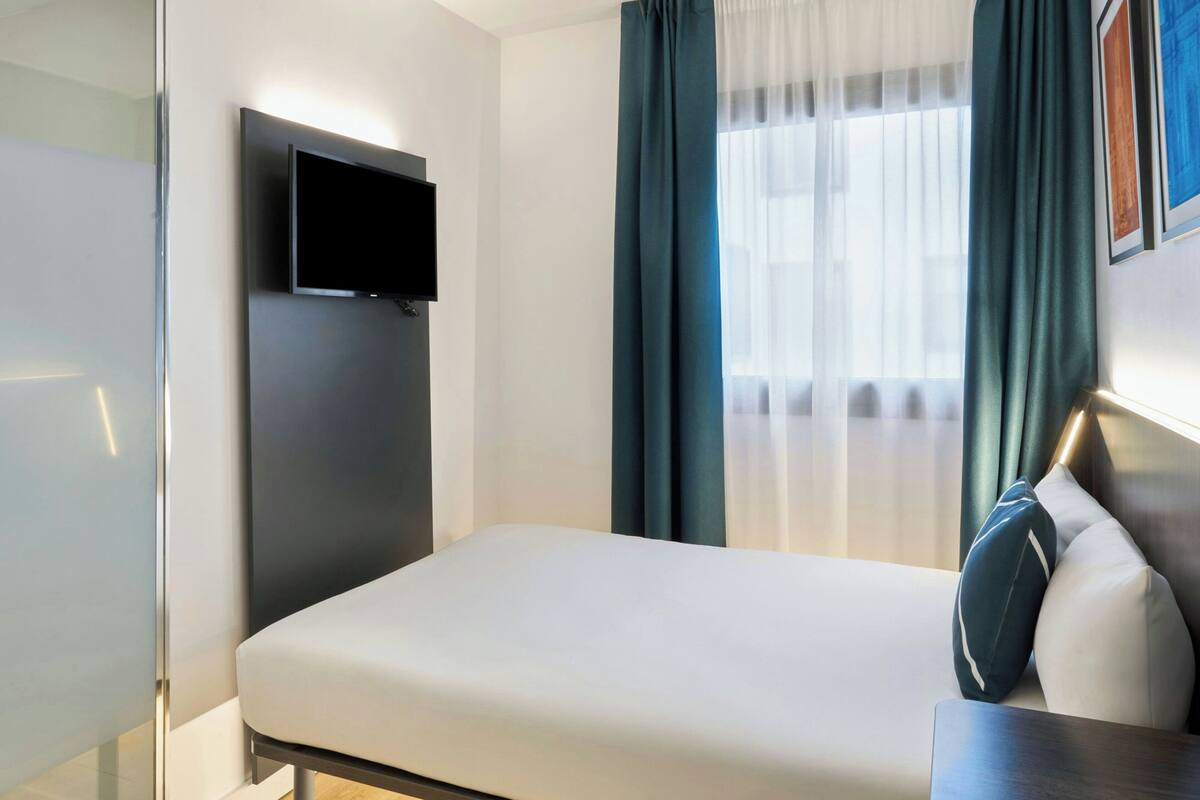 A hotel room with a bed, a mounted TV, a desk with a lamp, and a window with curtains.