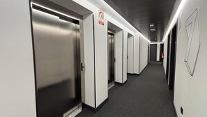 Un couloir avec plusieurs ascenseurs et portes, un extincteur et un panneau indiquant qu’il est interdit de fumer.