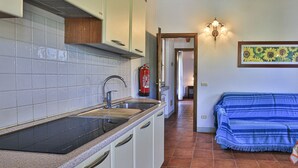 A kitchen with white cabinets, a black countertop, and a tiled backsplash. There is a fire extinguisher mounted on the wall, a sink, and a hallway leading to a room with a blue-striped sofa. - Perone, two-room apartment in a villa in Marina di Campo, with equipped garden, covered parking spac (Campo nell'Elba)