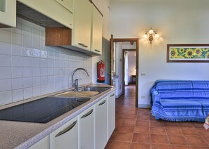 A kitchen with white cabinets, a black countertop, and a tiled backsplash. There is a fire extinguisher mounted on the wall, a sink, and a hallway leading to a room with a blue-striped sofa.