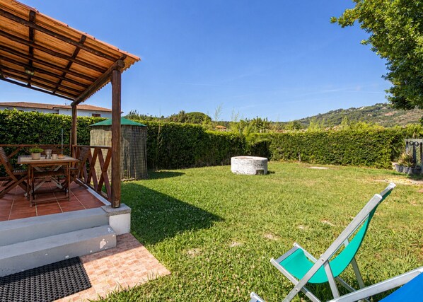 Un patio con mesa y sillas de madera, una jardinera de piedra y un jardín con césped, seto y vistas a colinas.
