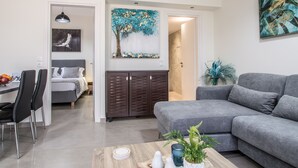 A modern living room with a grey sofa, a dining area with a table and chairs, and a bed with a grey headboard. - Luxury Katsoulakis Apartments (Alikianos)