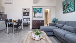 A modern living room with a grey sofa, a dining area with a table and chairs, and a kitchen visible in the background. - Luxury Katsoulakis Apartments (Alikianos)