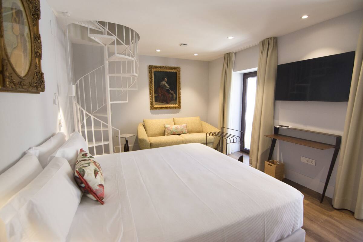 A hotel room with a large bed, a sofa, a TV, a painting, and a spiral staircase.