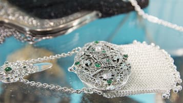 A silver necklace with intricate designs and green gemstones, displayed on a reflective surface.