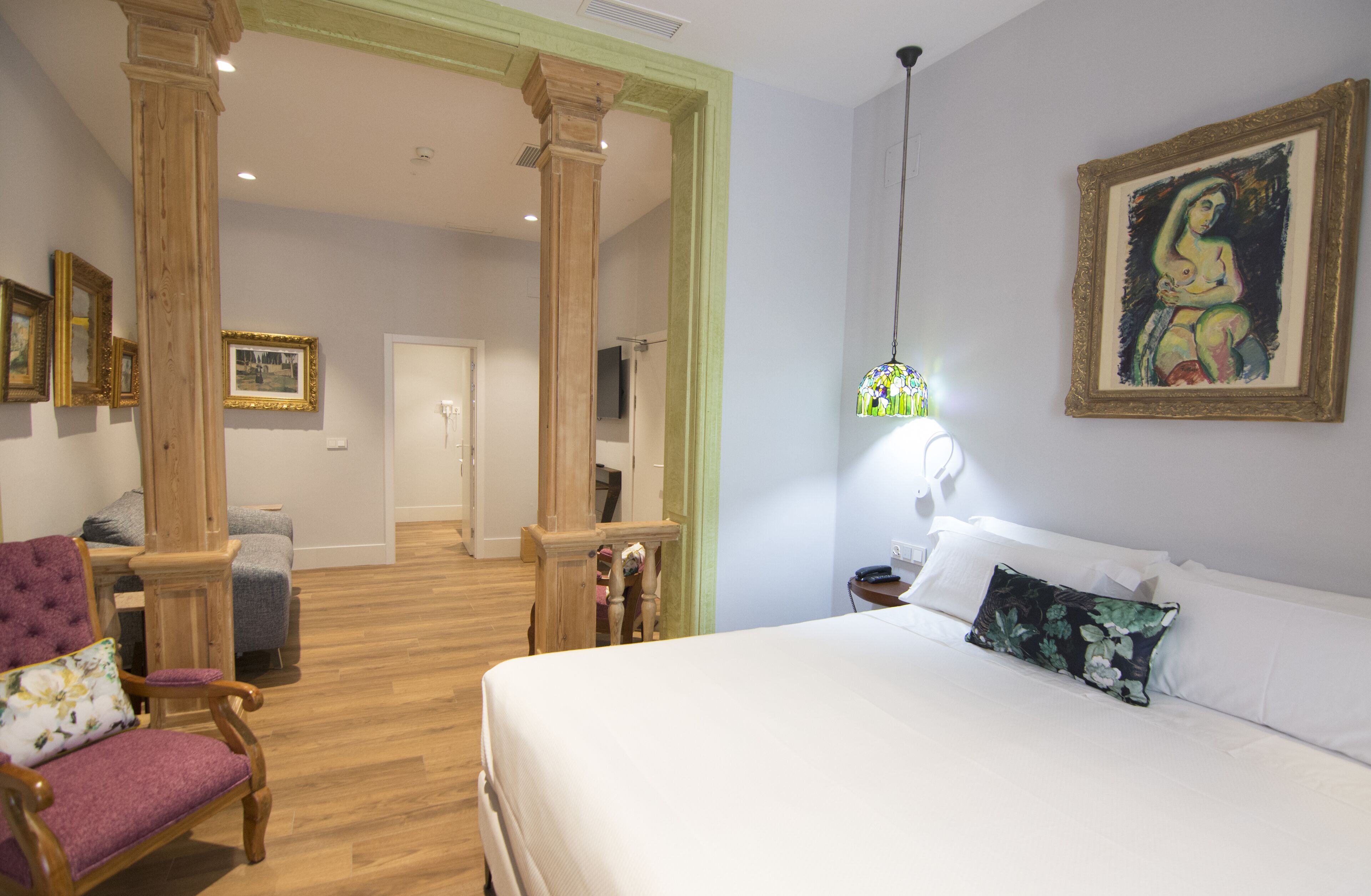 A hotel room with a large bed, a wooden chair, a desk, and a mirror. There is a painting on the wall and a door leading to another room.