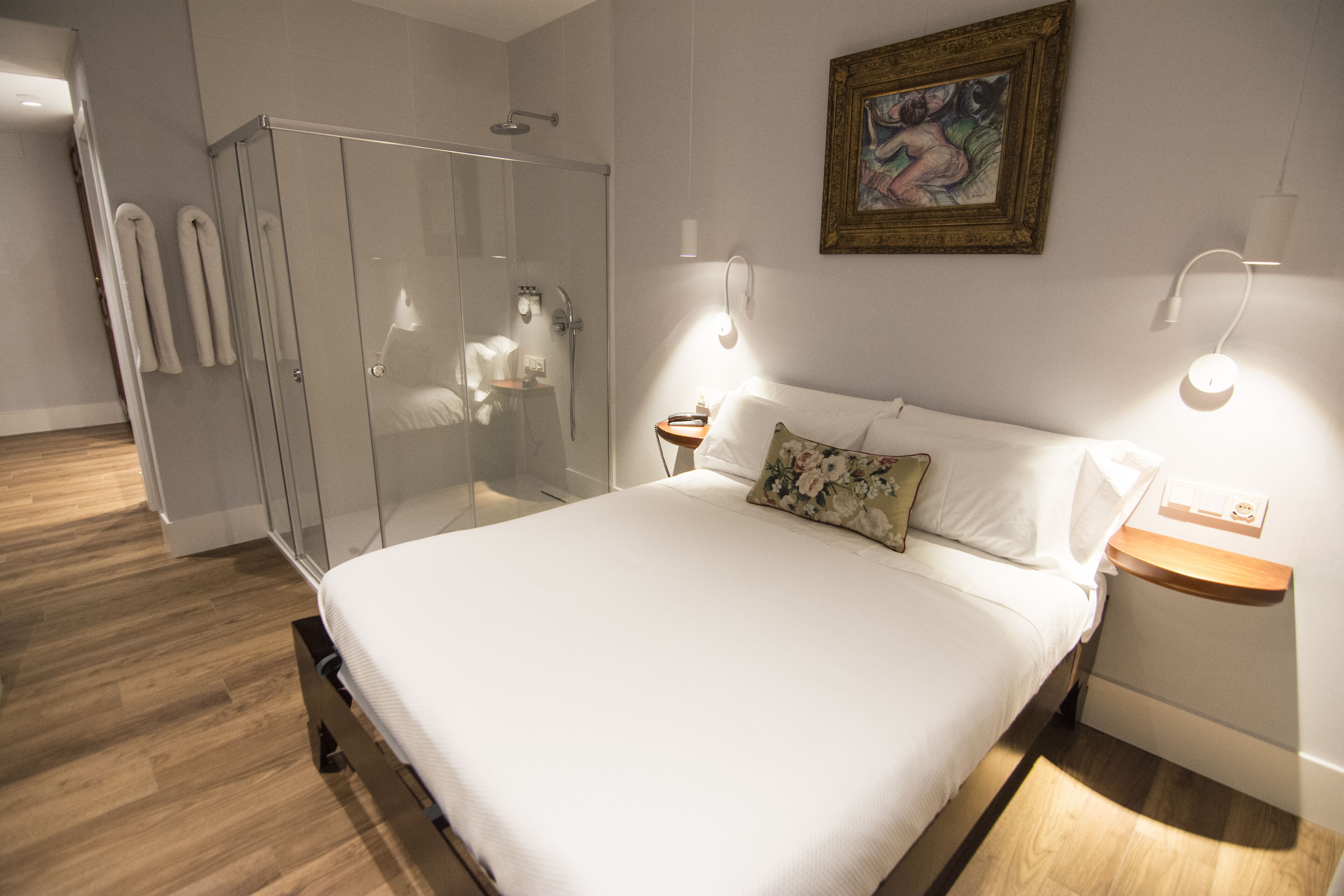 A bedroom with a bed, a glass shower enclosure, a nightstand, and a wall-mounted light fixture.