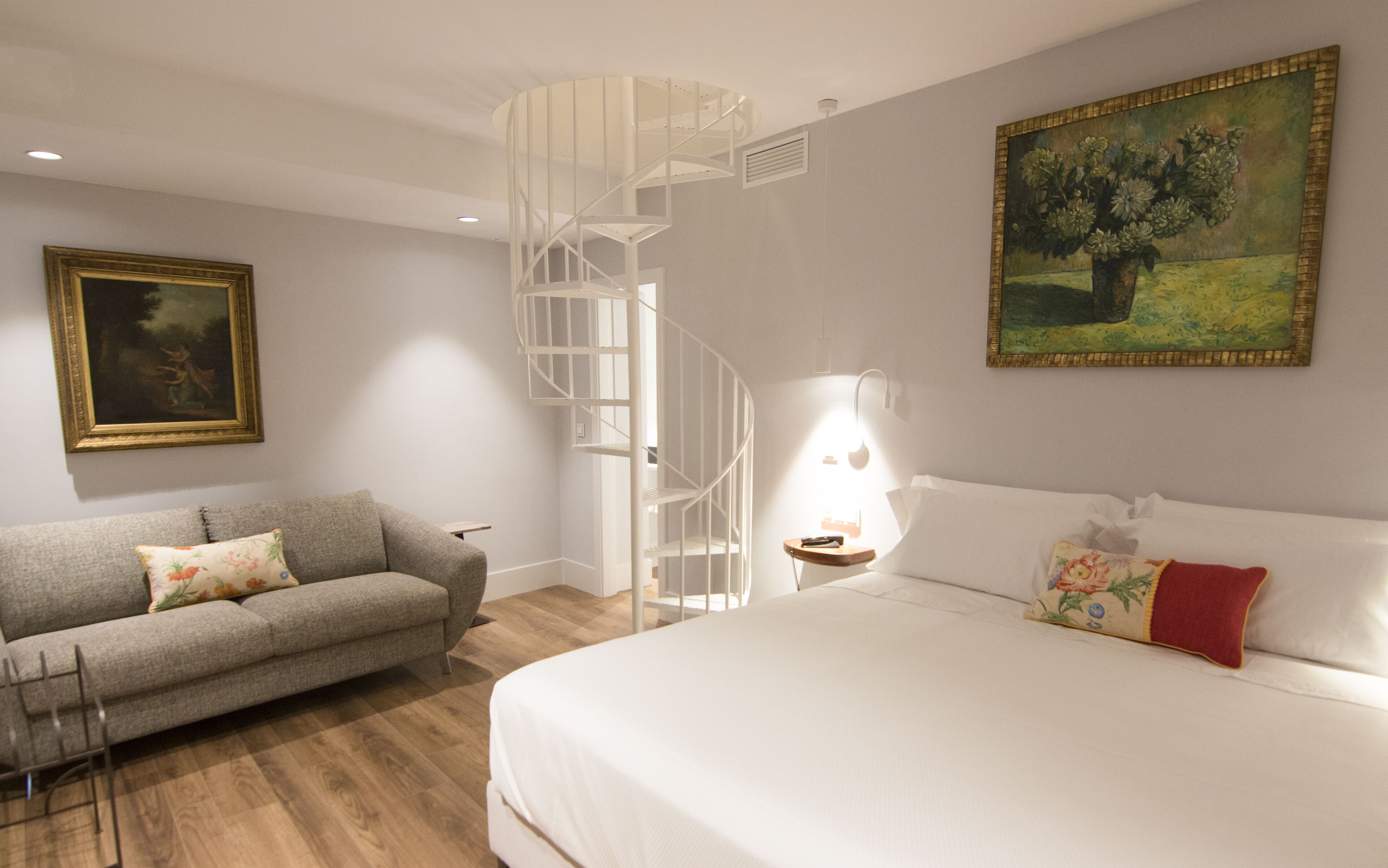 Un dormitorio moderno con una cama grande, un sofá gris, una escalera de caracol, un cuadro y una mesita de noche con una lámpara.