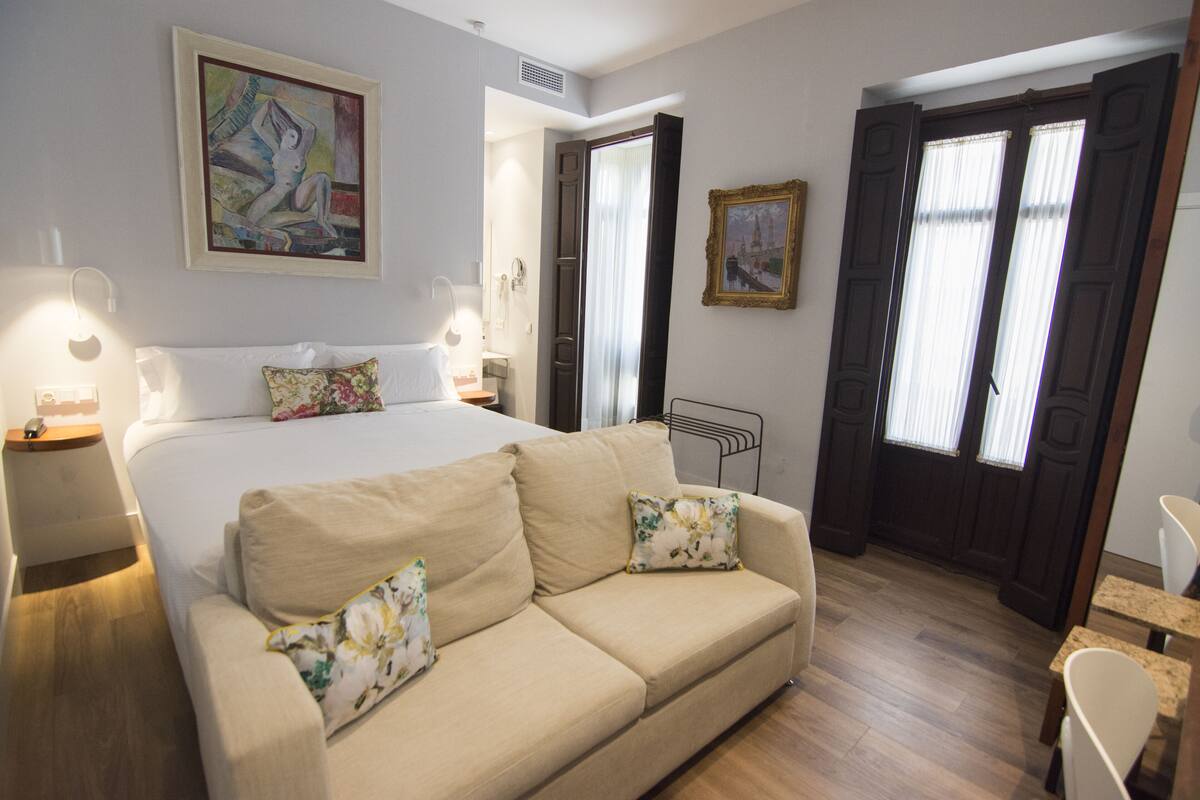 A hotel room with a bed, a sofa, a desk, and a chair. There are paintings on the walls and a window with curtains.