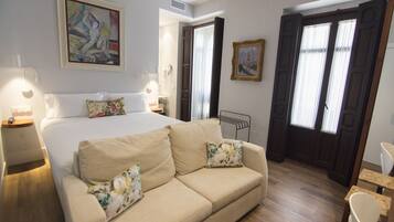 Habitació d'hotel amb llit, un sofà beix, una taula petita i dues cadires. Hi ha quadres a les parets i una porta amb cortines.