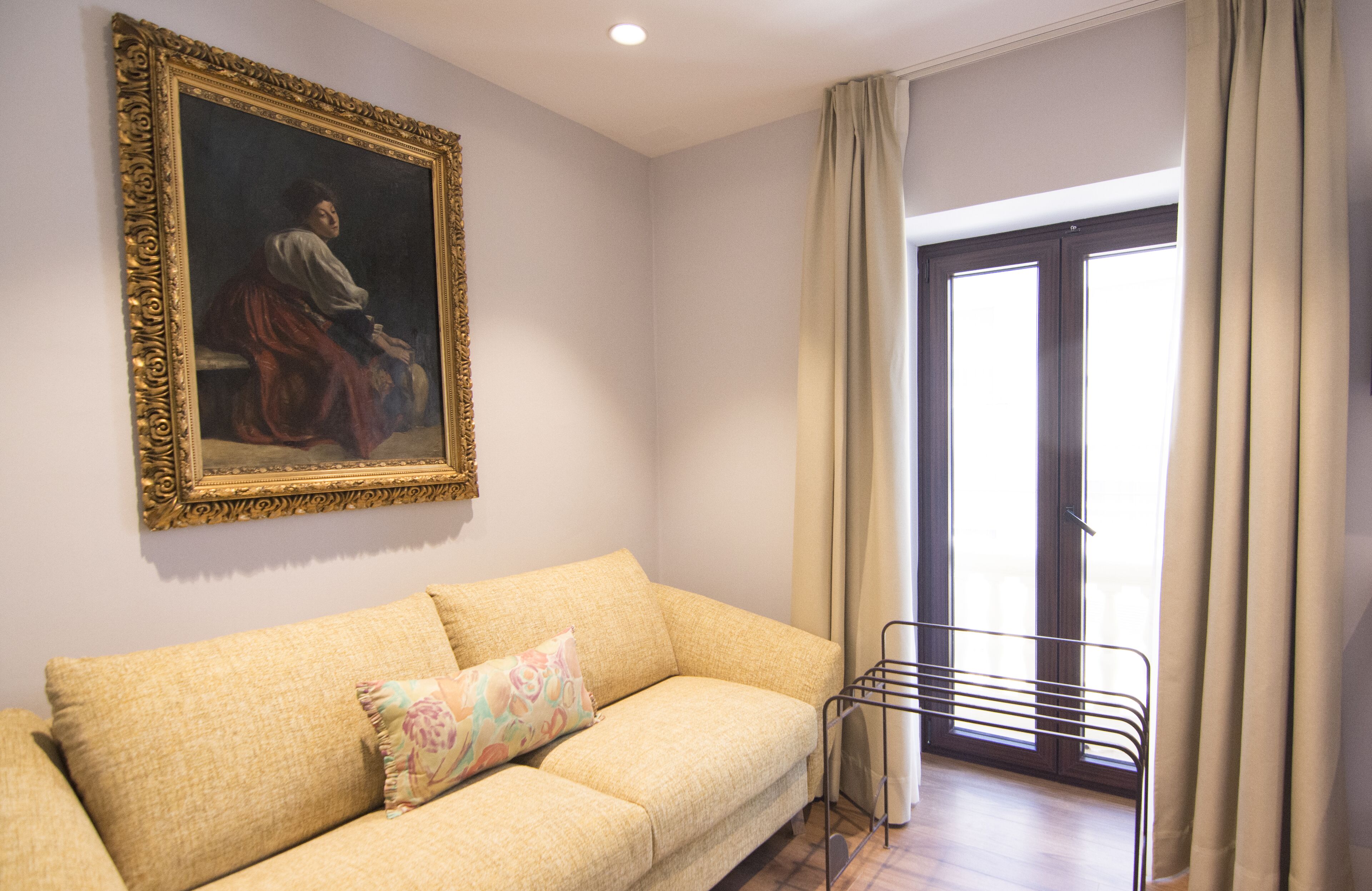 Una habitación con un sofá color beige, un cuadro enmarcado en la pared, una ventana con cortinas y un tendedero de metal.
