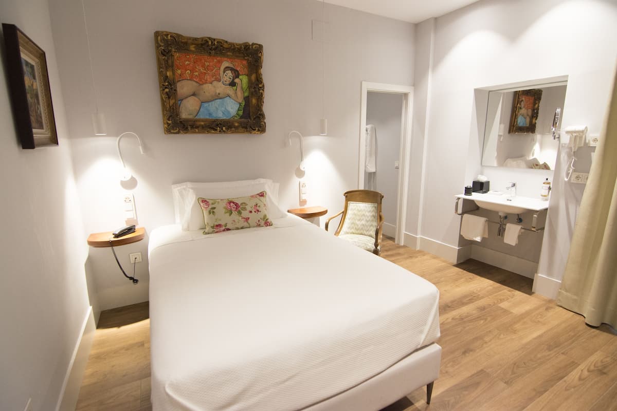 A bedroom with a bed, bedside tables, a chair, a painting, and a bathroom with a sink and mirror.
