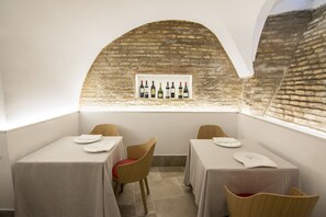 A dining area with two tables, each with a white tablecloth and a red chair. A brick wall and a window with a framed picture of wine bottles are visible. - Hotel Oleum (Andujar)