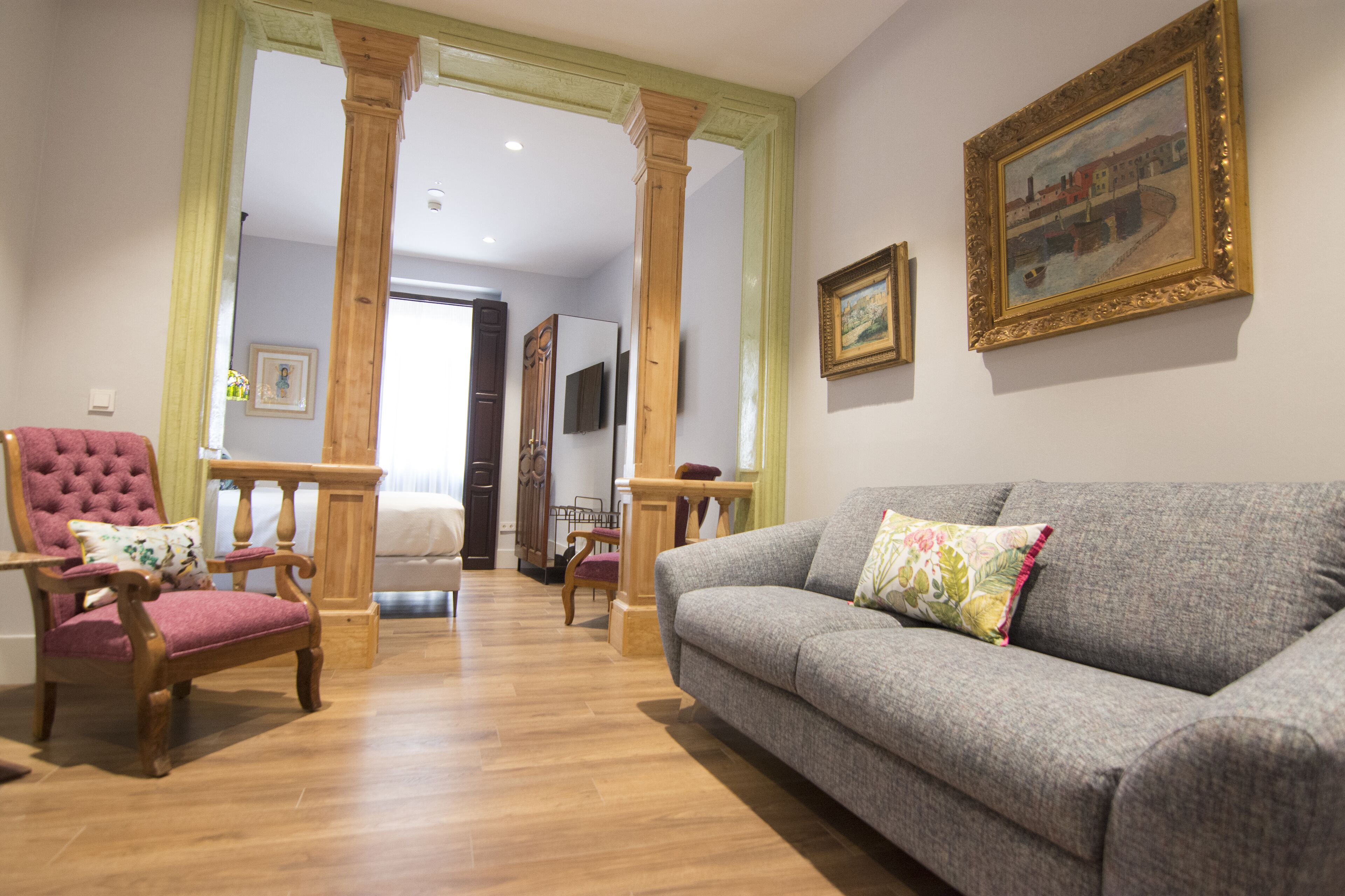 A living room with a grey sofa, a wooden armchair, and a wooden chair with a cushion. There is a painting on the wall and a wooden column in the background.