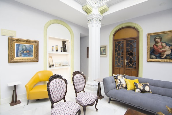 Una habitación con un sillón amarillo, un sofá gris y dos sillas estampadas. Se observan obras de arte enmarcadas en las paredes y una puerta de madera con un panel de vidrio decorativo.