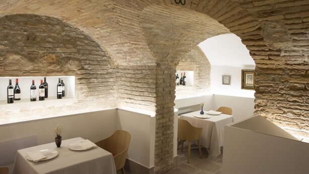 A stone-walled interior with tables and chairs, wine bottles on display, and a hanging light fixture. - Hotel Oleum (Andujar)