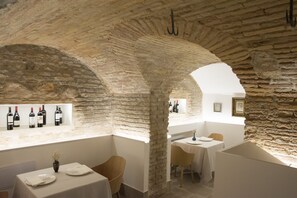 Interior de paredes de piedra con mesas y sillas, botellas de vino exhibidas y una lámpara colgante.