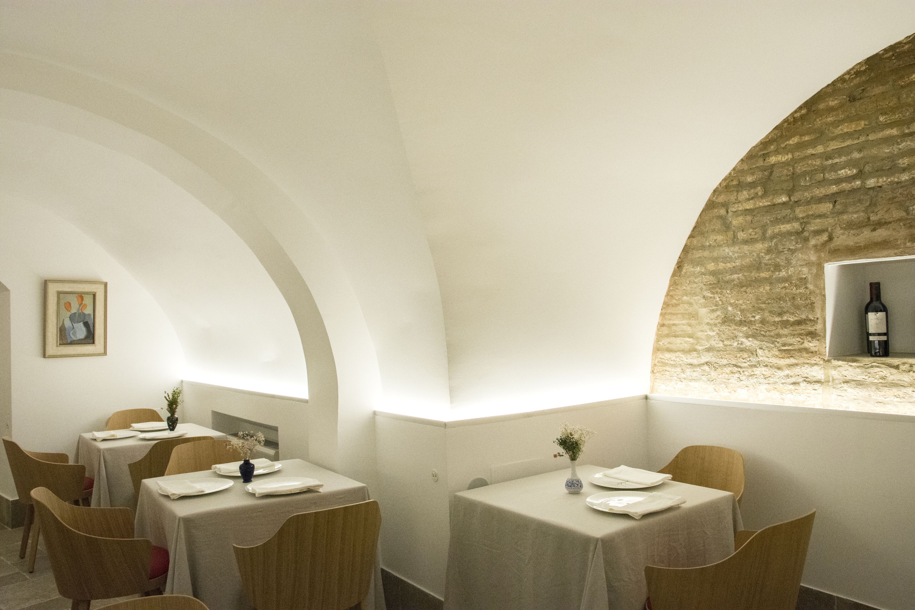 A restaurant with tables set for dining, a wine bottle on a shelf, and a framed picture on the wall.