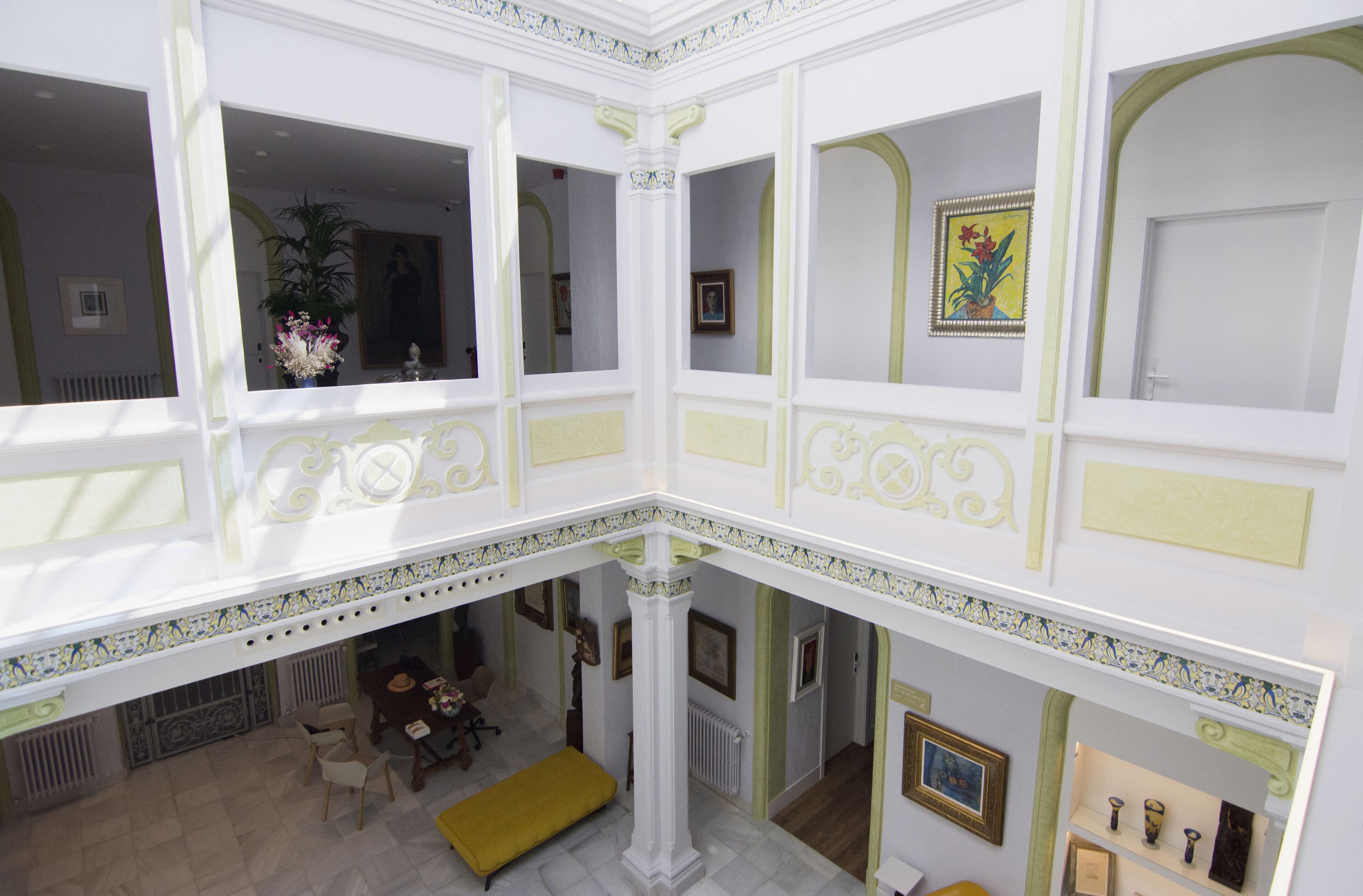 A spacious atrium with high ceilings, decorative moldings, and a central column. There are multiple seating areas with chairs and tables, and artwork is displayed on the walls.