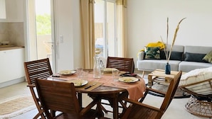 A dining area with a table set for four, surrounded by wooden chairs. A sofa and a small table with a vase of dried plants are visible in the background.