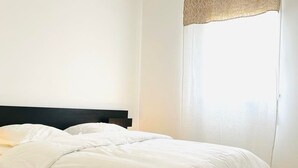 A neatly made bed with white linens in a room with a wooden floor and a plain white wall.