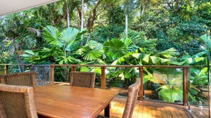 Un espacio exterior para comer con mesa de madera y sillas de mimbre, rodeado de exuberante follaje tropical.
