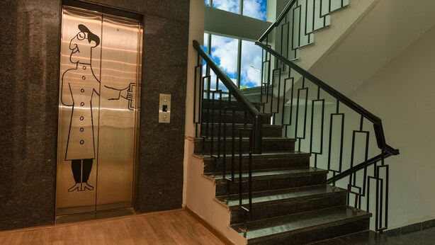 An elevator with a cartoon character, a staircase with black railings, and a view of the sky through the windows. - Hotel Olive Saffron (New Delhi)