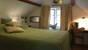 Ein Schlafzimmer mit einem Bett, einem Sessel, einem Schreibtisch, einem Spiegel und einem Fenster mit Vorhängen.