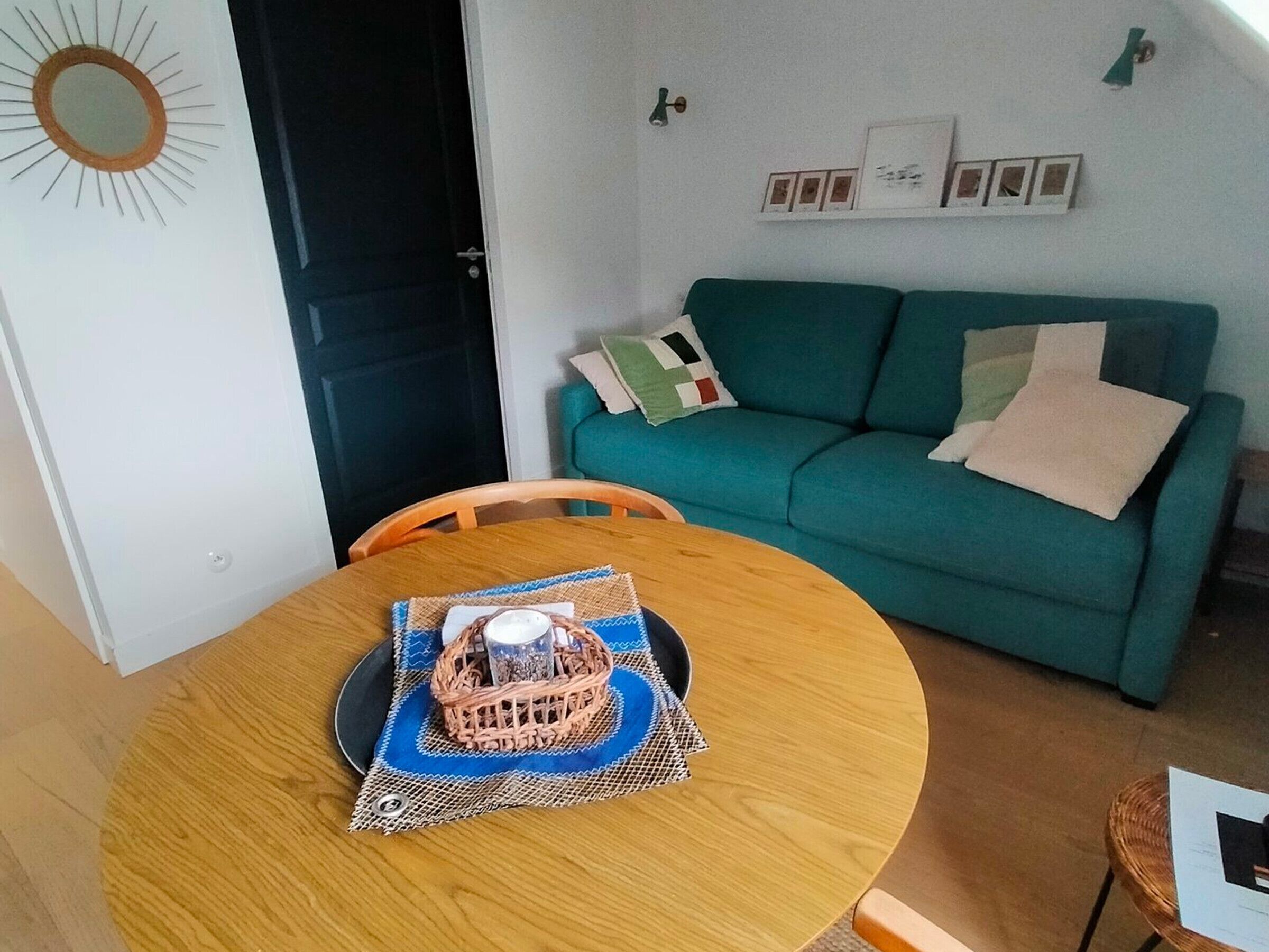 Un coin repas avec une table ronde en bois, un panier tressé contenant un récipient en verre et un canapé turquoise agrémenté de coussins.