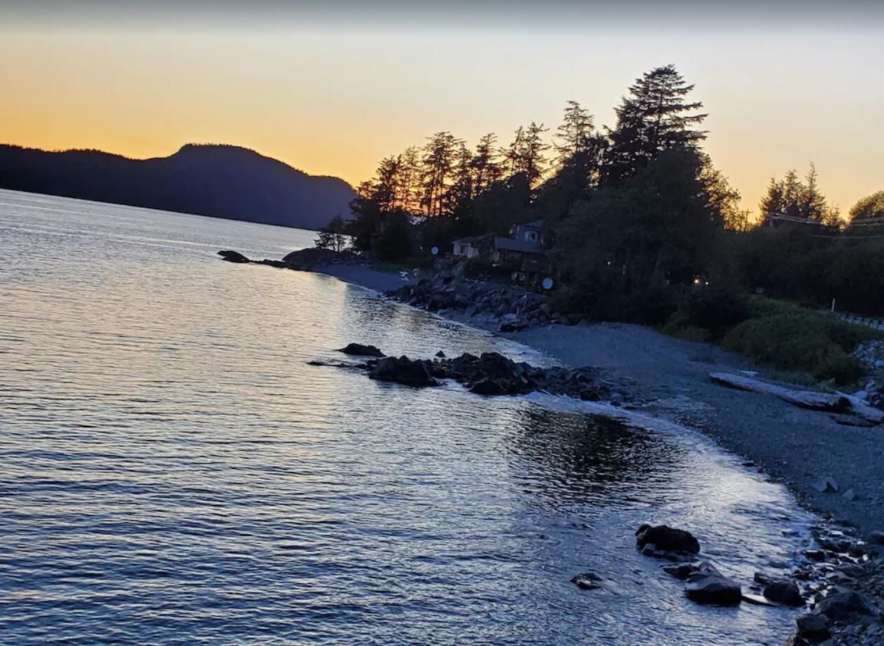 A coastal scene with calm water, a rocky shoreline, and dense forested hills in the background.