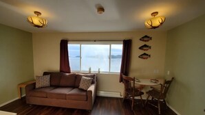 A living room with a brown sofa, a dining table with chairs, a window with curtains, and a view of the water. - {Forget-Me-Not Inn} Alaskan Waterfront Vacation Rental (Eagle Suite) (Sitka)