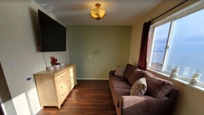 A living room with a flat-screen TV mounted on the wall, a wooden cabinet, a brown sofa with patterned cushions, and a large window with a view. - {Forget-Me-Not Inn} Alaskan Waterfront Vacation Rental (Eagle Suite) (Sitka)