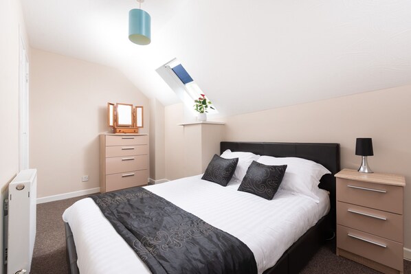 A bedroom with a bed, bedside table, lamp, dresser, and a window with a plant. - An apartment that sleeps 6 guests in 3 bedrooms (Ilfracombe)