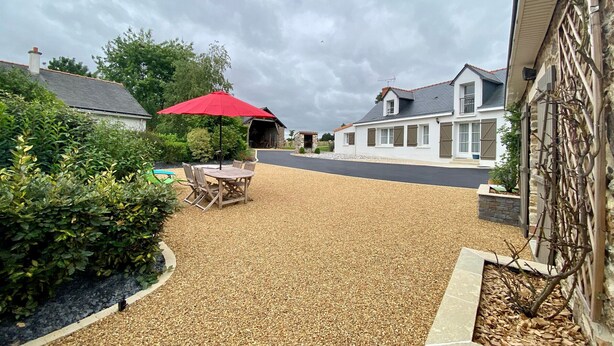 A residential driveway with a gravel surface, a red umbrella, and a wooden table with chairs.